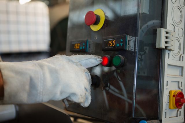 Hand wearing safety gloves pressing control button on industrial machinery in factory. Useful for illustrating manufacturing processes, industrial automation, safety protocols, and factory operations.