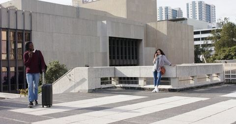 Diverse travelers pulling suitcase, leaning on rooftop railing and speaking on phones, urban plaza