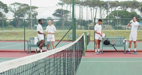Diverse male tennis players resting at net on outdoor hard court checking rackets