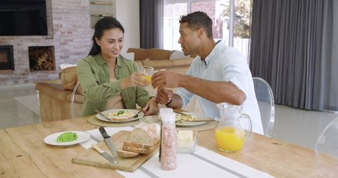 Multicultural couple clinking orange juice glasses during cozy breakfast at wooden table