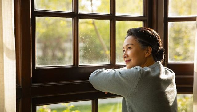 Senior asian woman leaning on wooden window rail gazing at sunlit garden, peaceful