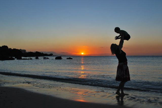 A mother is joyfully lifting her child into the air at the beach during sunset. The silhouettes of the pair against the colorful horizon create a heartwarming and picturesque scene. Ideal for themes of parenting, family bonding, summertime fun, and outdoor activity. Perfect for use in family-oriented advertisements, travel brochures, or inspirational content about motherhood and joy.