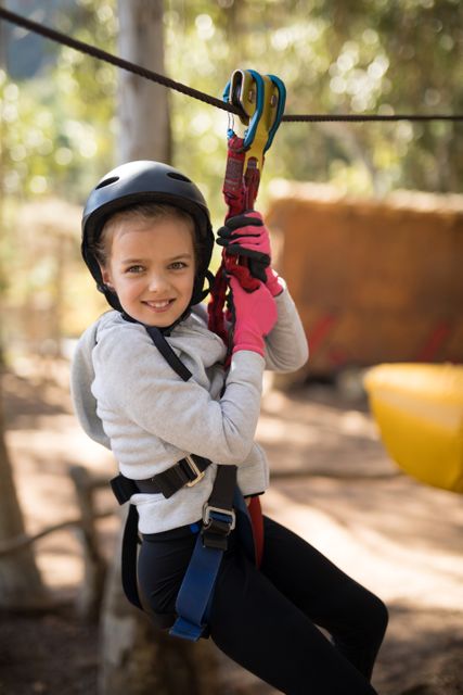 Young girl smiling while enjoying a zip line adventure in a forested area. She is wearing a helmet, safety harness, and gloves, indicating a focus on safety while having fun. This image is perfect for promoting outdoor activities, adventure parks, family outings, and children's recreational programs.
