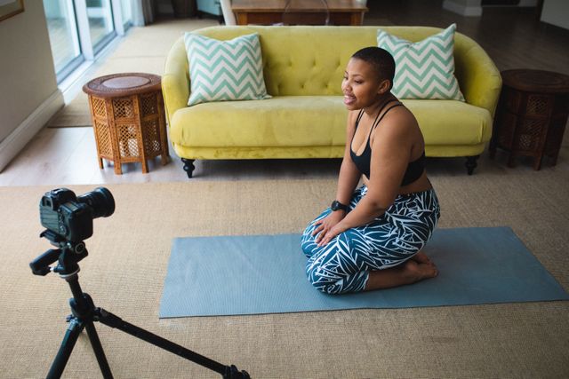 African American woman recording a yoga video at home, sitting on a yoga mat in a living room. She is using a digital camera on a tripod. Ideal for content related to fitness, healthy lifestyle, home workouts, yoga tutorials, and vlogging.