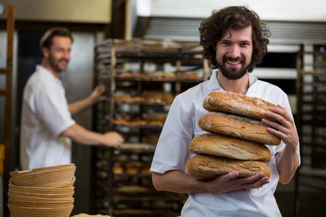Male baker smiling while holding stack of freshly baked bread loaves in a commercial kitchen. Ideal for bakery advertisements, culinary blogs, food industry promotions, and articles on professional baking.
