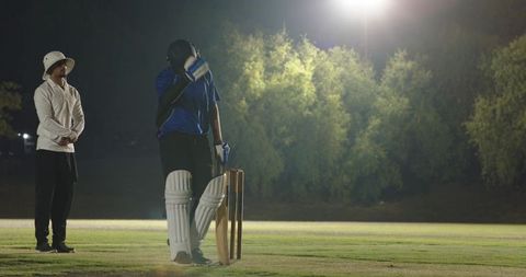 Night cricket batter adjusting helmet and pads at crease under floodlights with umpire