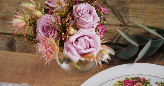 A bouquet of delicate pink roses and complementary flowers is arranged in a clear vase on a rustic wooden table, with a vintage floral plate nearby. The arrangement exudes a charming and romantic atmosphere, perfect for a cozy home setting or a special occasion.