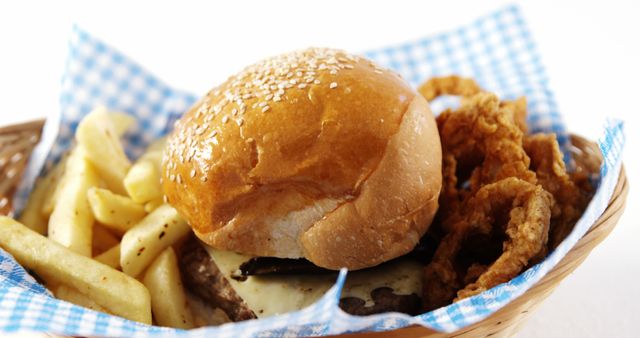 A cheeseburger with sesame seeds on the bun is accompanied by crispy onion rings and seasoned fries, served in a basket with a checkered liner. This appetizing meal is a classic example of American fast food cuisine, often enjoyed for its comforting and savory flavors.