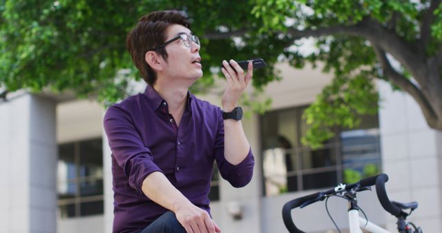 Asian businessman sits outdoors in an urban setting, using a smartphone to perform a voice search. He sits on a bench next to a bicycle, suggesting an active lifestyle, casual yet professional look in his purple shirt. Useful for themes on modern communication, technology in daily life, professionals on the go, and urban living.
