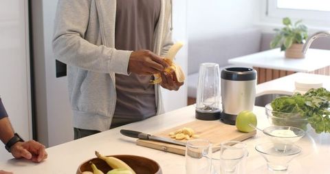 Diverse men preparing healthy smoothie at modern kitchen island, peeling banana