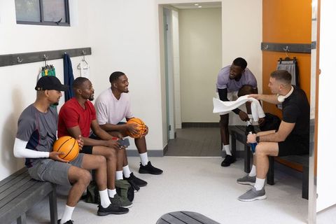 Multiracial basketball teammates sitting, chatting, passing towel in locker room postgame