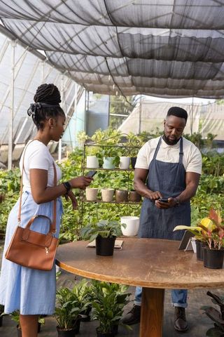 African american vendor and customer paying with smartphones at greenhouse nursery checkout
