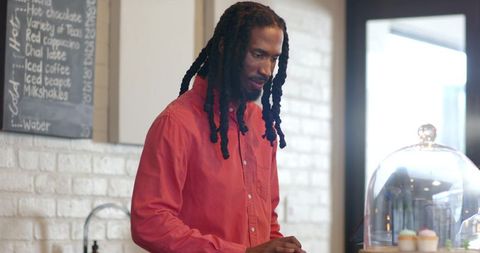 Barista with dreadlocks using tablet at café counter with cupcakes and chalkboard menu