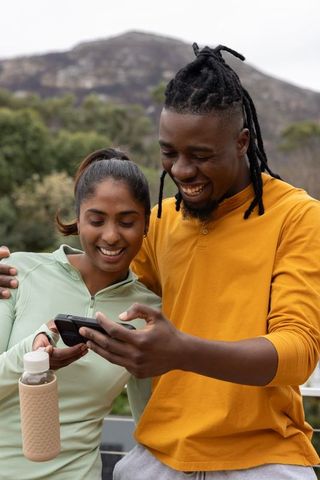 Smiling diverse couple sharing smartphone on balcony with reusable water bottle