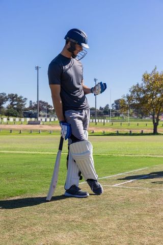 Cricketer inspecting batting glove at crease on sunny pitch with protective gear