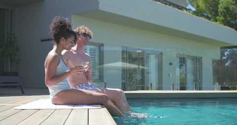 Relaxing couple sipping cocktails by modern backyard pool, dipping feet in clear water