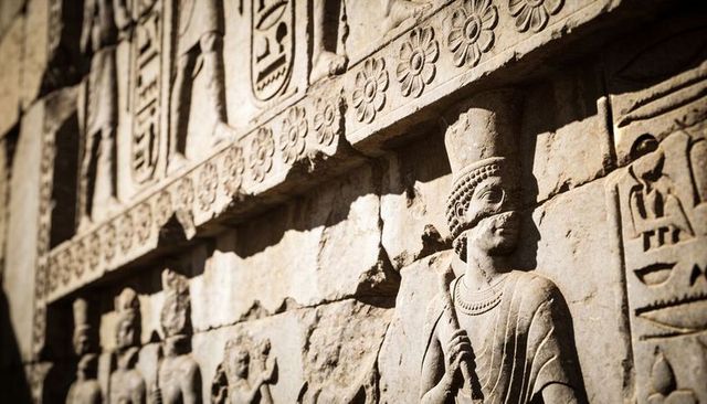 Ancient stone relief dignitary holding staff with crown and beaded necklace on wall
