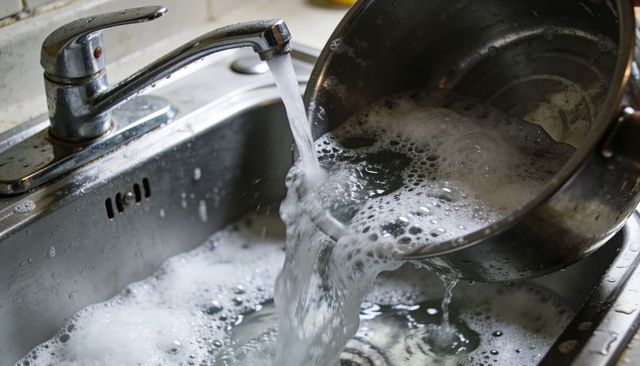 Pouring soapy water from stainless steel pot into kitchen sink with running faucet, foam