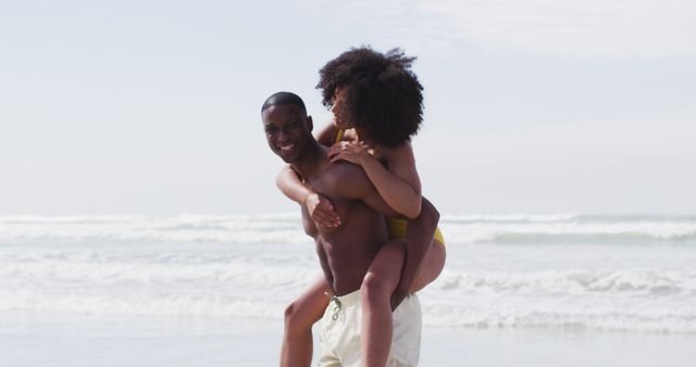 Smiling african american couple playing at the beach, man carrying woman piggyback. healthy outdoor leisure time by the sea.