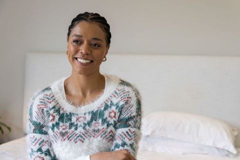 Smiling african american woman wearing cozy fuzzy sweater sitting on bed in neutral bedroom
