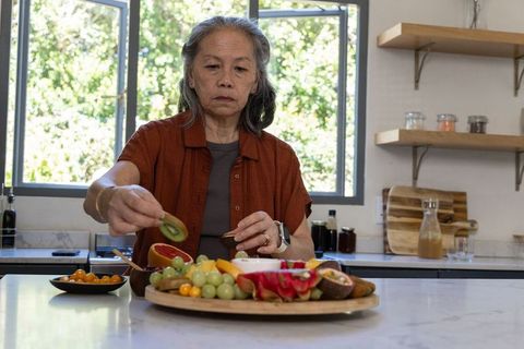 Senior asian woman arranging colorful wooden fruit platter on sunlit marble kitchen island