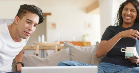 African american couple laughing on sofa watching laptop and sharing coffee moment
