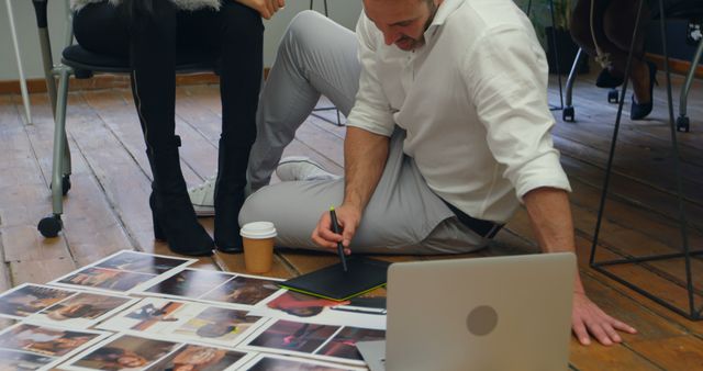 Caucasian man reviews photos in an office setting. He's engaged in selecting images for a project, surrounded by prints and a laptop.