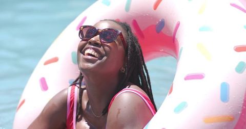 Smiling african woman relaxing on pink donut pool float wearing cat-eye sunglasses summer