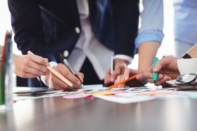 A close-up view of a creative team collaborating on a project. Several hands are seen holding pens and highlighters while discussing documents on a table in a modern office. Ideal for concepts related to teamwork, business meetings, creative processes, and project planning.