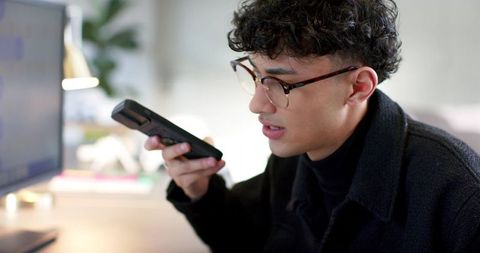Young man recording voice message on smartphone at modern home desk with monitor and lamp