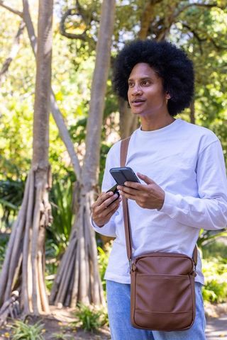 African american man checking phone and key fob in sunlit park with leather crossbody bag