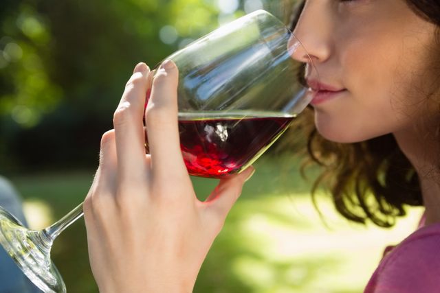 This image shows a woman enjoying a glass of red wine in a park, highlighting relaxation and leisure in a natural setting. Ideal for use in lifestyle blogs, wine advertisements, outdoor event promotions, and articles about relaxation and enjoying nature.