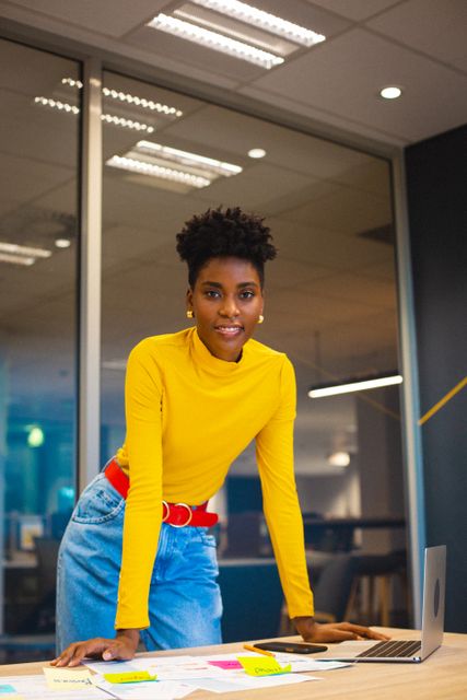 Young African American businesswoman standing confidently in a modern office environment, smiling at the camera. She is wearing a yellow top and blue jeans with a red belt, exuding professionalism and approachability. Ideal for use in business, corporate, and professional contexts, showcasing diversity, leadership, and modern work culture.