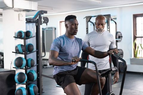 African american senior trainer coaching client on assault bike in modern gym