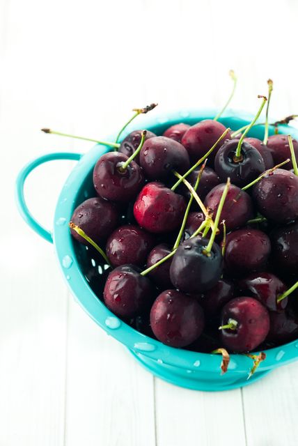 Bright, fresh, red cherries fill a vibrant blue colander, set against a clean white background. The cherries are glistening with water droplets, signaling that they have been recently washed. The contrast between the rich red of the cherries and the vibrant blue of the colander creates a visually appealing scene. This can be used for food blogs, healthy eating campaigns, summer recipes, or kitchen decor promotions.