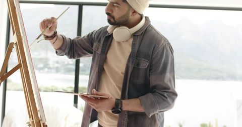 Young indian artist painting in bright waterfront studio wearing beanie and headphones
