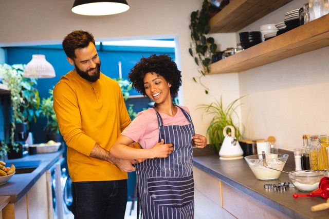 Bearded biracial young man assisting girlfriend in wearing apron while cooking in kitchen. Copy space, unaltered, lifestyle, love, togetherness, tying, afro hair, preparation and home concept.