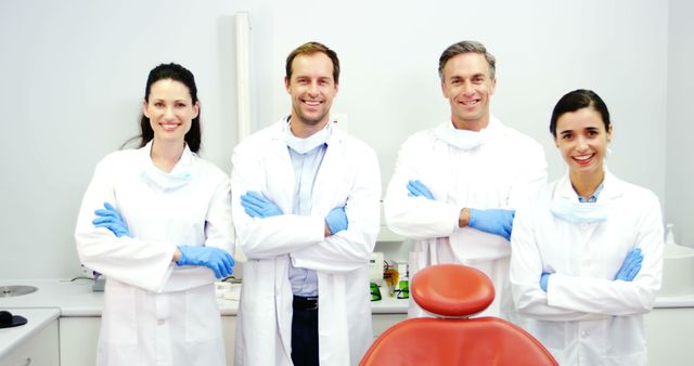 A diverse group of medical professionals, dentists or doctors, are smiling confidently in a clinical setting, with copy space. Their white coats, gloves, and the dental chair in the background suggest a healthcare environment focused on patient care.