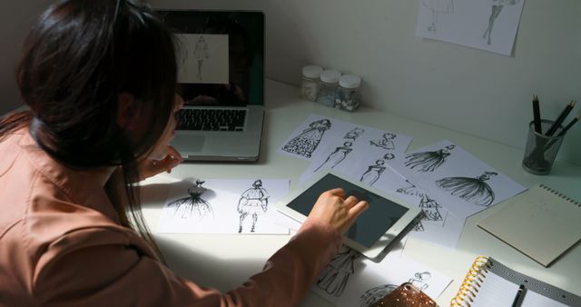 Caucasian female fashion designer looking at sketches using laptop and tablet at desk in studio. Communication, fashion, design, fashion studio, work and creative business, unaltered.