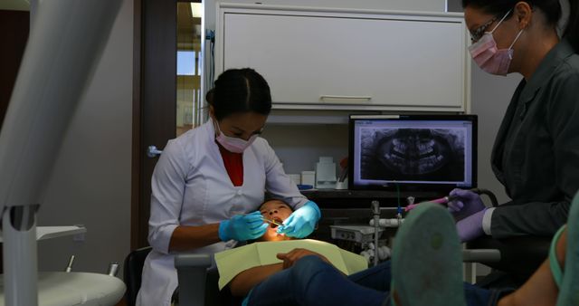 Pediatric dentist wearing white coat examining teeth of child sitting in dental chair in a modern dental office. Dental assistant sitting nearby, observing the procedure. Dental X-ray displayed on computer screen. Perfect for illustrating themes of pediatric dentistry, oral healthcare services, dental examinations, and hygiene practices in a clinical environment.