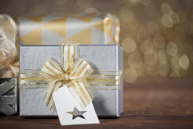 Close-up of beautifully wrapped gift boxes with gold ribbon and a tag on a wooden table, set against a festive bokeh background. Ideal for use in holiday promotions, Christmas advertisements, gift wrapping tutorials, and festive greeting cards.
