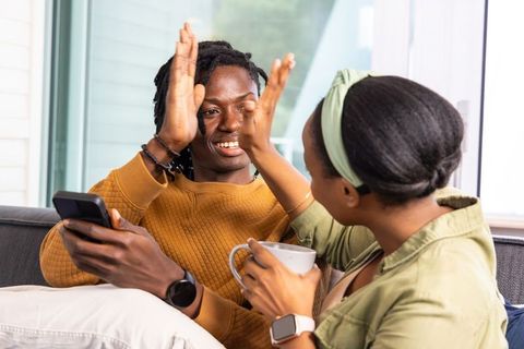 Young african american couple celebrating with high five on cozy sofa holding phone and mug