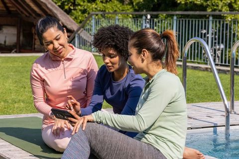 Multicultural women sharing smartphone while relaxing on poolside yoga mat in activewear