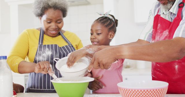 African American grandmother, granddaughter, and another family member baking together in modern kitchen. Grandmother and young girl sifting flour, smiling and enjoying the activity. Ideal for content showcasing family bonding, multi-generational activities, home cooking, and cultural traditions.