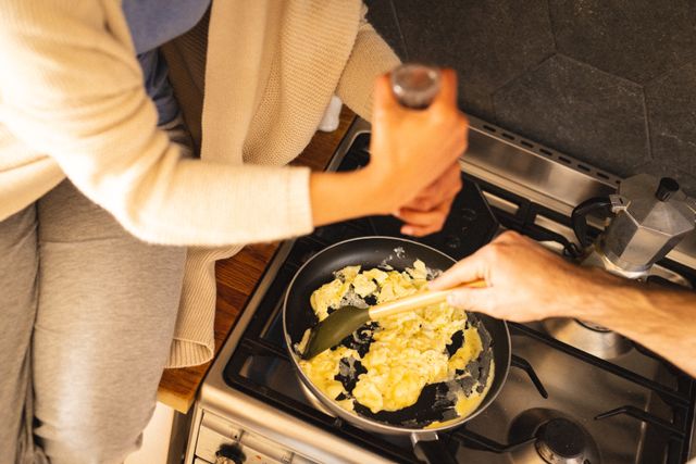 Hands of biracial man making scrambled eggs in cooking pan and girlfriend spraying pepper. Unaltered, lifestyle, love, togetherness, cooking, food, kitchen and home concept.
