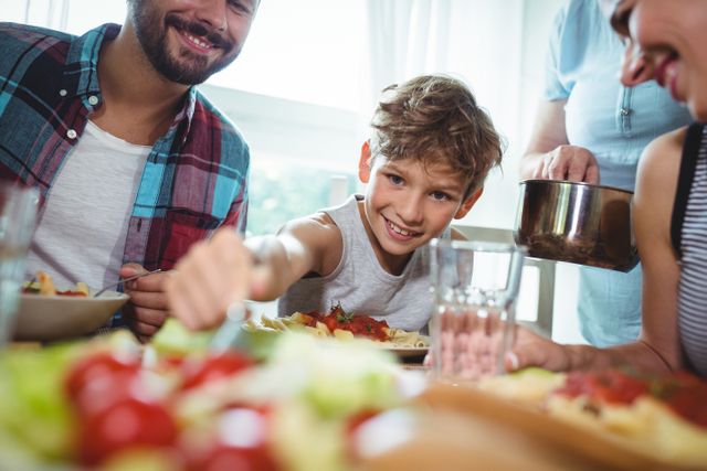 Boy enjoying a meal with his family at home, sitting at a dining table with food in the background. Ideal for use in advertisements promoting family values, healthy eating, parenting articles, and lifestyle blogs focused on family life.