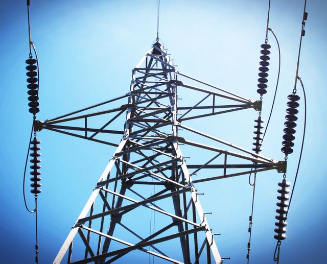 High angle view of a high voltage transmission tower with electric power lines extending against a clear blue sky. Suitable for use in articles about energy, power industry, electrical engineering, and infrastructure projects.