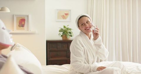Smiling woman wearing white bathrobe enjoying skincare routine on bed in cozy bedroom