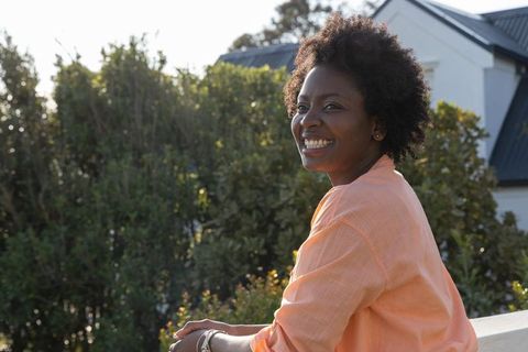 African american woman leaning on balcony railing smiling, looking right in sunlit garden
