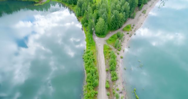 Aerial view captures a lush green landscape with a dirt path leading through trees between two bodies of water, reflecting the sky. The scene emphasizes the tranquility and beauty of natural environments.